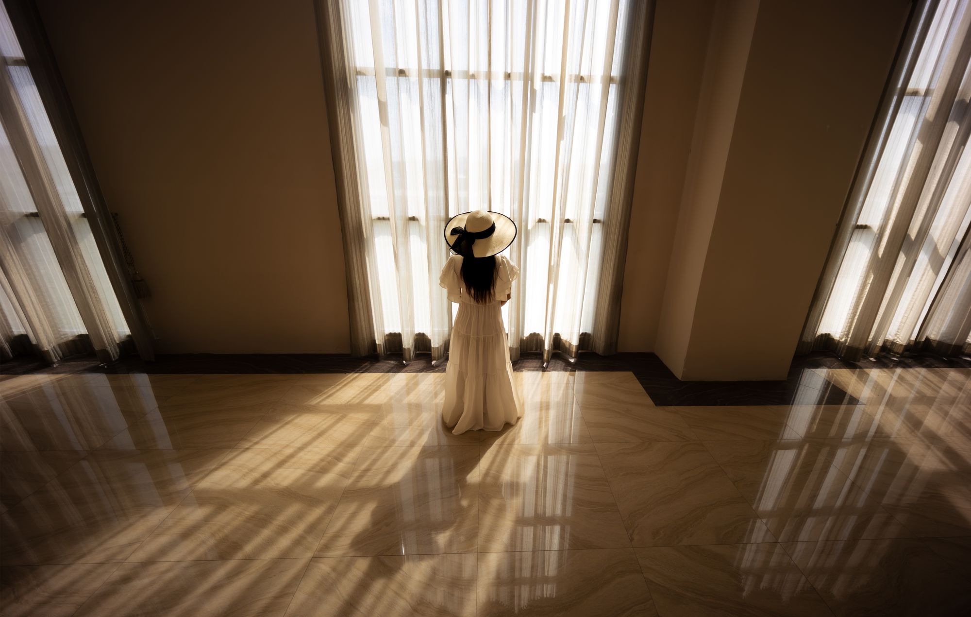 A woman in an ivory dress and sun hat before tall curtained windows